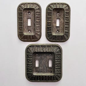 May include: Three rectangular light switch plates with a dark metallic finish and a textured, brushed surface. The borders feature an intricate, raised pattern of small floral motifs and scrolling filigree details surrounding the central switch openings.