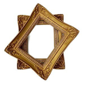 May include: Two gold-coloured picture frames with elaborate scrollwork are arranged diagonally against a white backdrop. The frames have a classic, vintage design. The frames are empty, ready for photos or artwork.