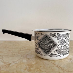 May include: White enamel saucepan with a black handle and a lid. The exterior features a black and white fish pattern. The saucepan is designed for cooking and has a long, black handle for easy handling.