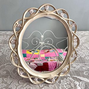 May include: A round, cream-colored mirror with a decorative floral frame. The frame features an openwork design with small, pink flowers. The mirror reflects a bed with a pink quilt and a white headboard, set against a light blue wall.