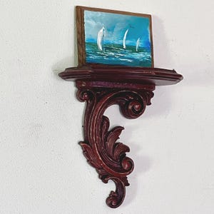 May include: A dark red, ornate wall shelf with a small painting of sailboats on the sea. The painting is framed in a light brown wood. The shelf has a curved front and decorative, scrolling support.