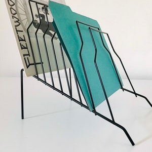 May include: A black metal record and file holder with a slanted design. The holder has vertical bars and supports a record album and a teal file folder. The album cover features the word "Fleetwood" and a black and white image.