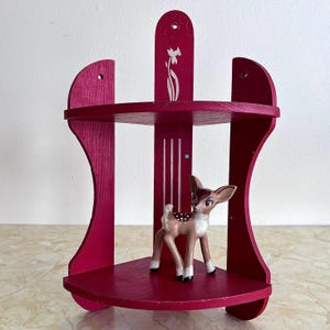 May include: A magenta corner shelf with two tiers and a decorative floral design. A small ceramic deer figurine with white spots and black hooves is displayed on the bottom shelf. The shelf is made of wood.