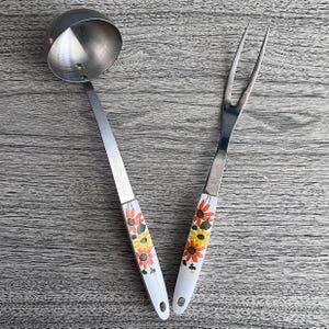May include: A stainless steel ladle and carving fork set. The handles are white with a floral design in orange, yellow, and green. The ladle has a round bowl. The carving fork has two tines.