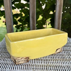 Peut inclure: Un pot de fleurs rectangulaire en céramique jaune avec une finition brillante. Le pot est surélevé par deux supports à texture bois. Le design est simple et moderne, adapté à l'intérieur ou à l'extérieur. Le pot mesure environ 30 cm de long.