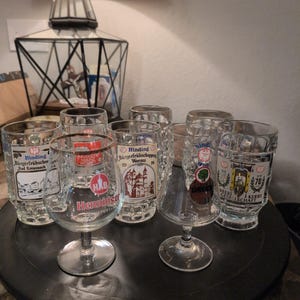 May include: A collection of vintage beer glasses with various printed designs and text. Some glasses are short and cylindrical, while others have stems. The text includes "Binding", "Bürgerfrühschopper", and "Henning". The glasses are arranged on a dark, round tray.