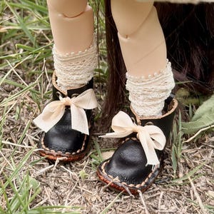 May include: A doll wearing a white dress with green stitching, beige lace-trimmed socks, and black leather-like shoes with cream ribbons. The doll carries a beige crocheted bag decorated with small, colorful red, yellow, and orange mushrooms.