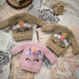 May include: Four miniature knit sweaters, each featuring a unicorn face design. The sweaters are in shades of tan and pink, with embroidered details including a golden horn, floral accents, and closed eyes. The sweaters are displayed on a vintage paper background.