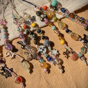 May include: A collection of beaded necklaces with various charms, including butterflies, shells, and stars. The necklaces feature a mix of colorful beads in shades of pink, blue, yellow, and green. Each necklace is unique, with different bead shapes and sizes.