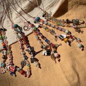 May include: A collection of beaded necklaces with various charms and colorful beads. The necklaces feature a variety of shapes, including flowers, butterflies, and hearts. The beads are in shades of pink, blue, green, and yellow.
