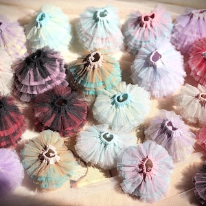 May include: A collection of colorful, layered tulle skirts in various pastel shades, including pink, purple, blue, and red. The skirts are arranged on a light-colored surface, showcasing their ruffled texture and delicate design. These skirts are suitable for dance or costume.