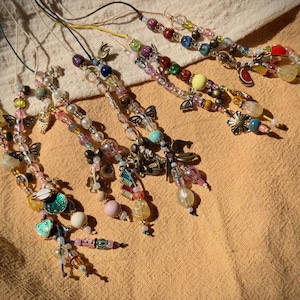 以下が含まれることがあります： さまざまな色と形をしたビーズチャームのコレクション。チャームは、パール、カラフルなガラスビーズ、装飾的な金属チャームなど、さまざまなビーズを組み合わせています。一部のチャームには、蝶、花、果物のデザインがあります。チャームは細いコードに紐付けられています。