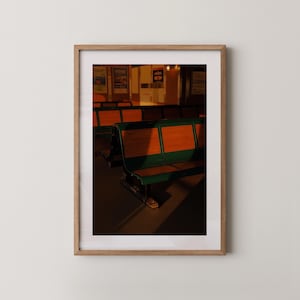 Könnte beinhalten: Gerahmtes Foto mit einer Reihe von Vintage-Bänken. Die Bänke haben grüne Metallrahmen und Sitze und Rückenlehnen aus Holz. Das Foto wird von warmem Sonnenlicht beleuchtet, das Schatten wirft. Der Hintergrund umfasst Poster und einen Türrahmen.