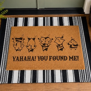 May include: A rectangular tan coir doormat featuring five black line-art illustrations of small forest creatures and the text "YAHAHA! YOU FOUND ME!" printed in black. The mat is layered over a black and white vertically striped rectangular rug.