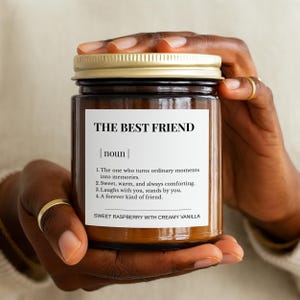 Puede incluir: Una vela en tarro de cristal marrón con tapa dorada. La etiqueta dice "THE BEST FRIEND" con una definición y la descripción de la fragancia "SWEET RASPBERRY WITH CREAMY VANILLA". La vela está sostenida por una persona.
