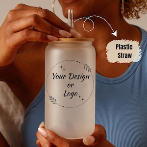 Może przedstawiać: Mrożony szklany kubek z bambusową pokrywką i przezroczystą plastikową słomką. Kubek ma okrągły wzór z napisem "Your Design or Logo". Projekt jest czarny. Osoba ma na sobie niebieski top.