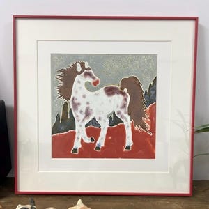 Op de afbeelding: Ingelijst kunstwerk met een wit paard met bruine vlekken en een bruine manen en staart. Het paard staat op een rode ondergrond tegen een grijze en blauwe achtergrond. Het kunstwerk is ingelijst in een rode lijst.