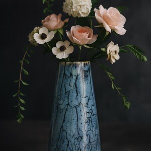 Könnte beinhalten: Eine blaue Keramikvase mit einem einzigartigen schwarzen Marmormuster enthält einen Blumenstrauß. Das Arrangement umfasst rosa Rosen, weiße Anemonen und eine Gruppe weißer Hortensien. Grünes Laubwerk ergänzt die Blumen in der Vase.