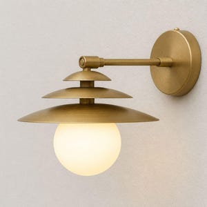 May include: A wall-mounted light fitting featuring a brushed brass finish with a circular backplate and a horizontal arm. The lamp includes three tiered, conical metal shades stacked above a single, spherical, frosted white glass globe bulb.