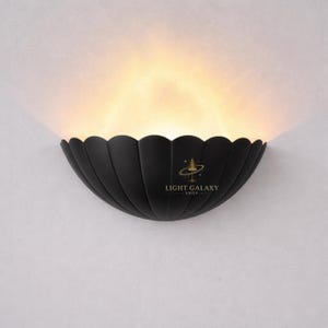 Puede incluir: Aplique de pared negro con un diseño festoneado, que emite una luz cálida y difusa. La lámpara tiene forma semicircular y presenta el texto "LIGHT GALAXY SHOP". El aplique está montado en una pared blanca.