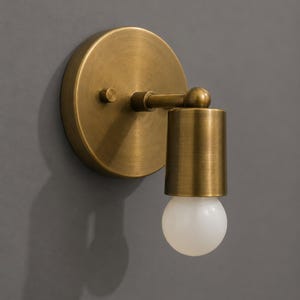 May include: A brass-toned wall sconce with a round backplate and a cylindrical shade. The sconce features a small arm and a spherical detail connecting the shade to the backplate. A white, round light bulb is visible.