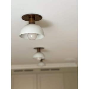 May include: White and brass-colored ceiling light fixtures. The fixtures have a rounded, bowl-shaped shade and a brass-colored base. The light bulbs are visible inside the shades. The image shows multiple fixtures, suggesting a home decor theme.