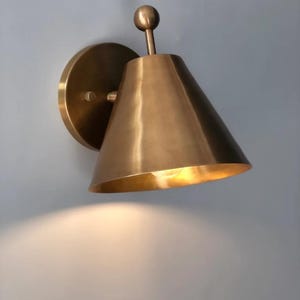 May include: A brass-toned wall sconce with a conical shade and a round backplate. The sconce has a small, spherical detail at the top and casts a warm light. The fixture is designed for indoor use and adds a touch of elegance.