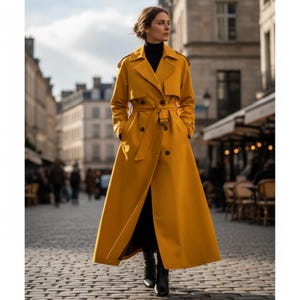 May include: A mustard yellow trench coat with a double-breasted design, a belt at the waist, and a long, flowing silhouette. The coat features a classic collar, button details, and pockets. The person is wearing black boots and a black turtleneck.
