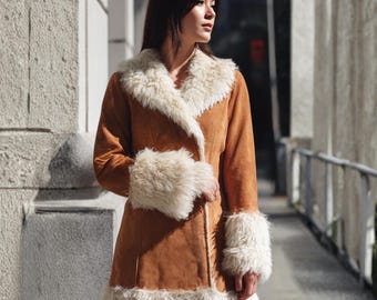 May include: A brown suede coat with cream-colored faux fur trim on the collar, cuffs, and hem. The coat has a double-breasted design and a mid-thigh length. The coat is worn outdoors.