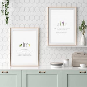 May include: Two framed art prints with gold frames are displayed on a white shelf. The left print features a cocktail illustration with text, while the right print shows a lavender and heart illustration with text. The prints are set against a white hexagon tile wall.