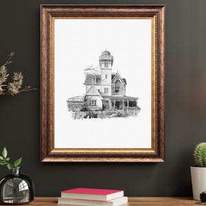 May include: A framed black and white architectural print of a Victorian-style house. The print is in a gold-toned frame and features a detailed illustration of the house with a tower, porch, and intricate details. The artwork is displayed on a wooden surface.
