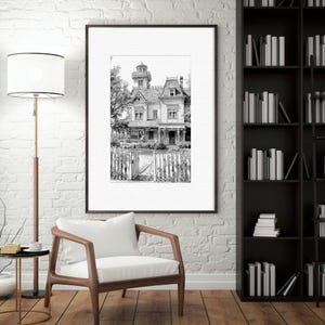 May include: Black and white framed print of a Victorian-style house with a picket fence and open gate. The artwork is displayed on a white brick wall, with a wooden chair and bookshelf in the foreground.