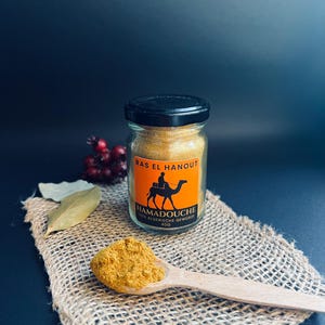 May include: A glass jar of Ras El Hanout spice with a black lid, orange label, and a camel illustration. A wooden spoon holds a portion of the yellow spice, placed on a burlap cloth. The background is a dark blue.