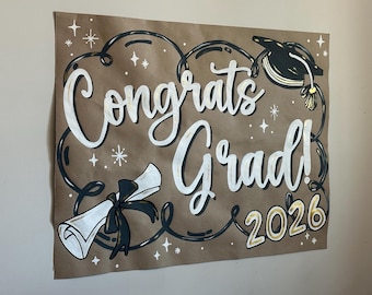 Könnte beinhalten: Ein braunes Papierschild mit den Worten "Congrats Grad!" in weißer Schrift, akzentuiert mit schwarzen Wirbeln, Sternen und einer Abschlusskappe. Das Jahr "2026" ist in Gold gehalten. Eine gerollte Urkunde mit schwarzem Band ist ebenfalls abgebildet.