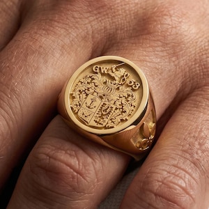 Puede incluir: Un anillo de sello dorado con un diseño de escudo grabado. El anillo presenta un emblema detallado con las iniciales "GW" y el año "'53". El anillo se lleva en un dedo, mostrando su acabado pulido y su intrincada artesanía.