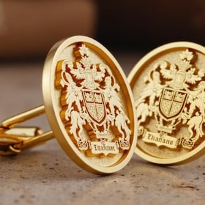 Puede incluir: Un par de gemelos dorados con un diseño de escudo de armas grabado. Los gemelos ovalados presentan un escudo detallado con un escudo, una corona y figuras de apoyo. La palabra "Thahana" está inscrita en un gemelo.