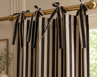 May include: A curtain panel featuring a vertical black and cream striped pattern. The curtain is suspended from a metallic gold rod by several fabric ties that are knotted at the top, creating a gathered effect along the upper edge of the fabric.