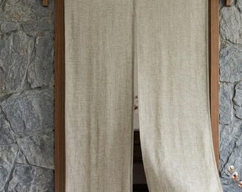 May include: A beige linen noren curtain featuring a split-panel design hanging from a wooden rod. The fabric has a visible woven texture and a neutral, light tan color. The curtain is suspended within a wooden door frame against a stone wall.