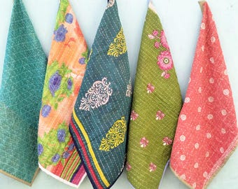May include: Five colorful, patterned fabric cloths hanging against a light blue wall. The cloths feature various designs, including floral and geometric patterns, in shades of green, orange, and pink. Each cloth is folded into a triangle shape.