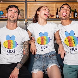 Può includere: Tre persone indossano magliette bianche con una grafica di un'ape colorata e la parola "you" in corsivo. Il design dell'ape presenta strisce arcobaleno. Le persone sorridono e ridono.