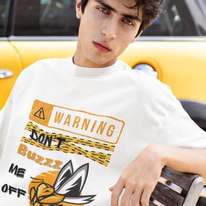 Può includere: T-shirt bianca con un design grafico con un segnale di avvertimento, nastro di precauzione e un'ape dei cartoni animati. Il testo sulla maglietta recita "WARNING DON'T BUZZ ME OFF". L'ape è gialla, nera e bianca.