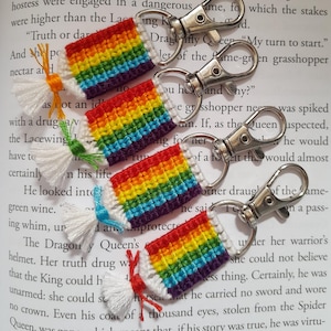Può includere: Quattro portachiavi arcobaleno fatti a mano con chiusure color argento. Ogni portachiavi presenta un design arcobaleno rettangolare all'uncinetto con un bordo bianco e un nappa in vari colori. I portachiavi sono esposti su una pagina di libro.