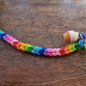 May include: A rainbow-braided bracelet featuring a cupcake charm and a green heart-shaped charm. The bracelet is made of small, woven rubber bands in various colours, including red, orange, yellow, green, blue, and purple.