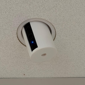 Puede incluir: Una cámara de seguridad cilíndrica blanca está instalada en un techo. La cámara tiene una lente negra y una luz azul. La cámara está montada en una abertura circular en el techo.