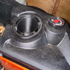 May include: Close-up of a black plastic component with a circular opening and a smaller, round cap. The cap has a red indicator. The component appears to be part of a larger machine or vehicle, with an orange section visible.