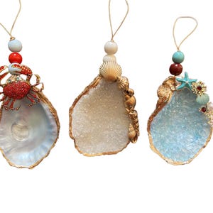 May include: Three decorative oyster shell ornaments. Each shell has a gold-painted edge and is embellished with various sea-themed elements. One features a red crab, another has seashells, and the third includes a starfish and other jewels.