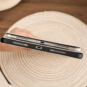 May include: A tablet device encased in a protective cover, featuring a black frame with visible speaker grilles and a central charging port. The device is positioned to show the side profile, highlighting the precise cutouts for hardware access.