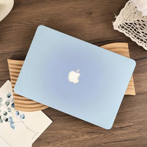 May include: A pale blue laptop with a white Apple logo rests on a wooden stand. The laptop's top is visible, showing its closed state. The background features a wooden table and decorative items.