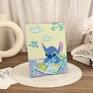 Puede incluir: Una portada de libro azul y amarillo con el personaje Stitch de Lilo & Stitch. Stitch está leyendo un libro con otros personajes de dibujos animados. La portada tiene un fondo a rayas amarillas y blancas con nubes y flores de dibujos animados.