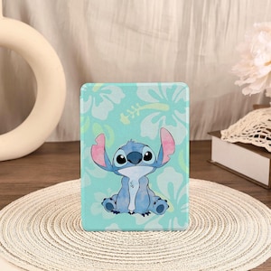 Puede incluir: Una funda para tableta azul claro con un personaje de dibujos animados, Stitch, con orejas grandes y ojos grandes. El fondo es azul claro con patrones florales blancos y verde claro. La funda es rectangular y está sobre una estera tejida.